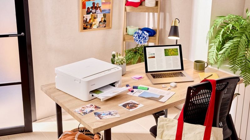 Stampante Canon su una scrivania mentre stampa documenti accanto a un laptop, con fotografie e articoli di cancelleria in uno spazio di lavoro domestico