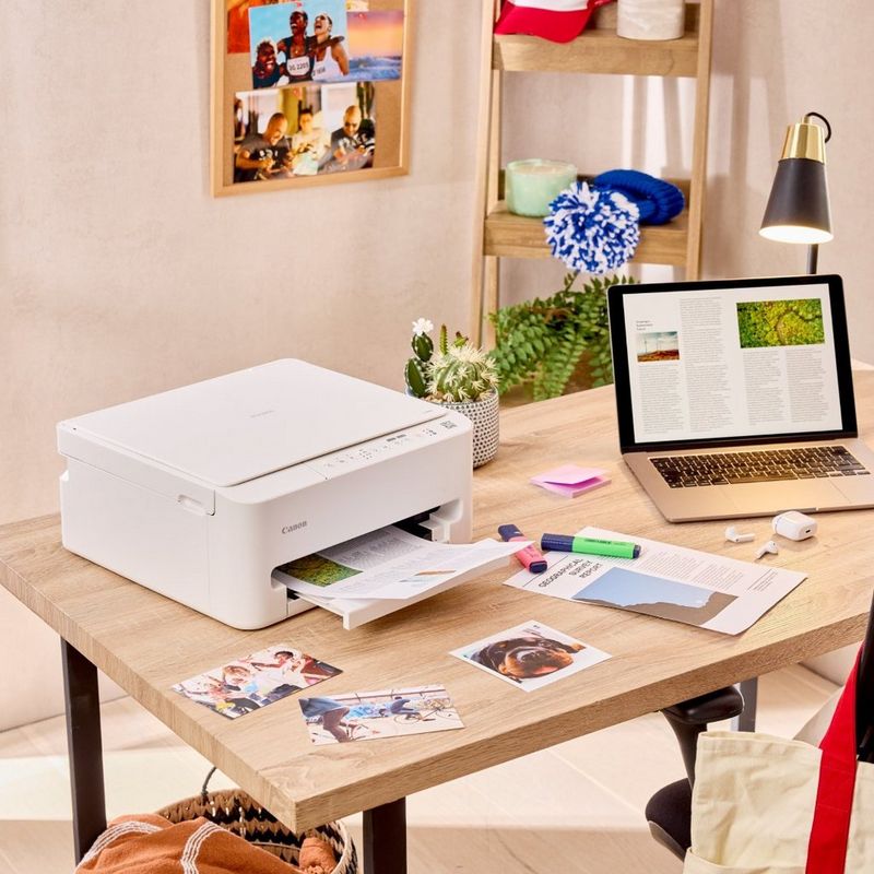 Stampante Canon su una scrivania mentre stampa documenti accanto a un laptop, con fotografie e articoli di cancelleria in uno spazio di lavoro domestico