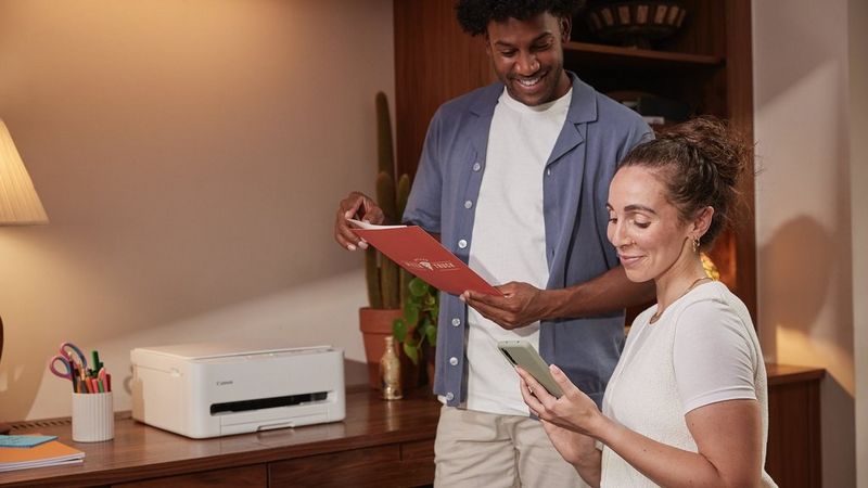 Two people using a Canon printer on a wooden desk in a warmly lit room