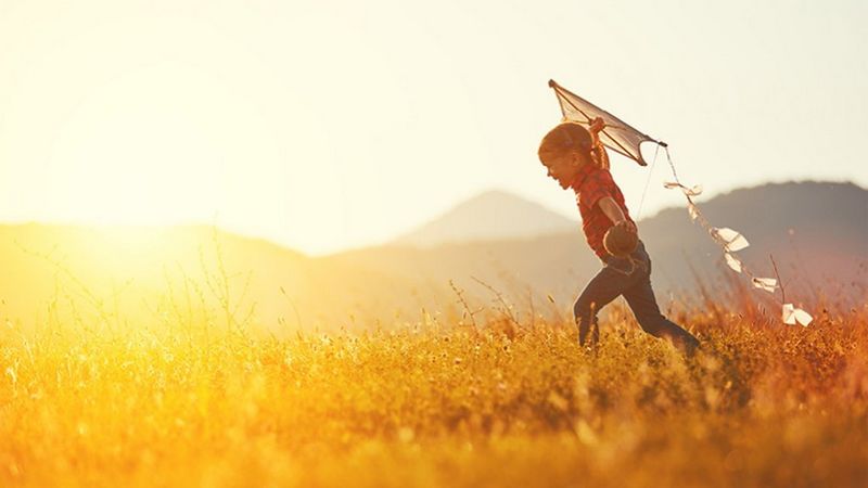 Enfant courant dans un champ ensoleillé tout en tenant un cerf-volant, avec des montagnes visibles en arrière-plan.