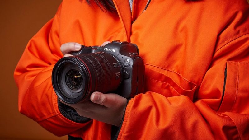 Photographe portant une veste orange vif et tenant un appareil photo hybride Canon EOS R6 équipé d'un objectif Canon.