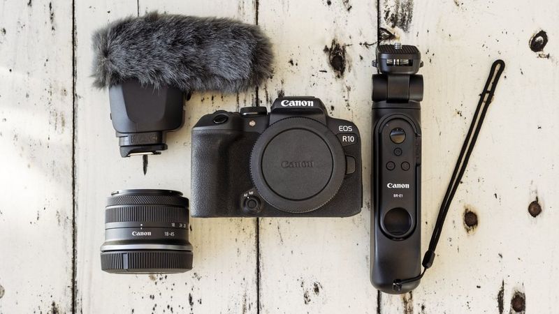 Boîtier de l'appareil photo Canon EOS R10 avec objectif, microphone et télécommande disposés sur une surface en bois blanc.