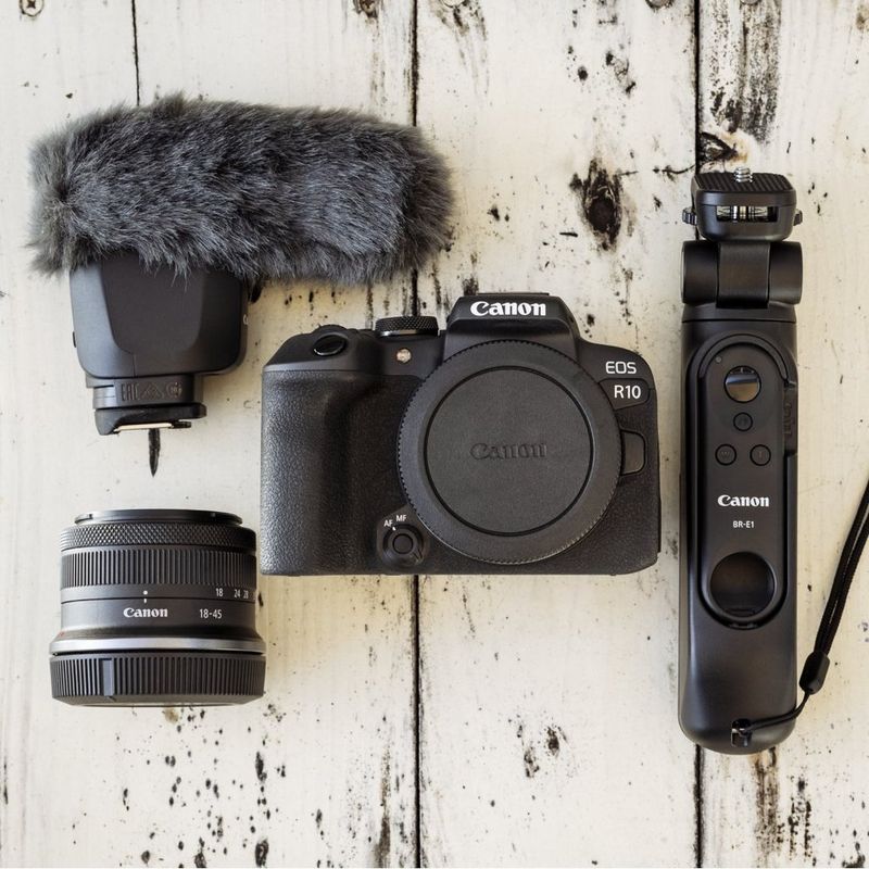 Boîtier de l'appareil photo Canon EOS R10 avec objectif, microphone et télécommande disposés sur une surface en bois blanc.