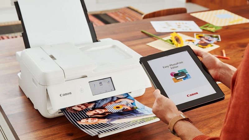 Easy-PhotoPrint Editor på en tablet ved siden af en Canon-printer, der printer et foto på et skrivebord.