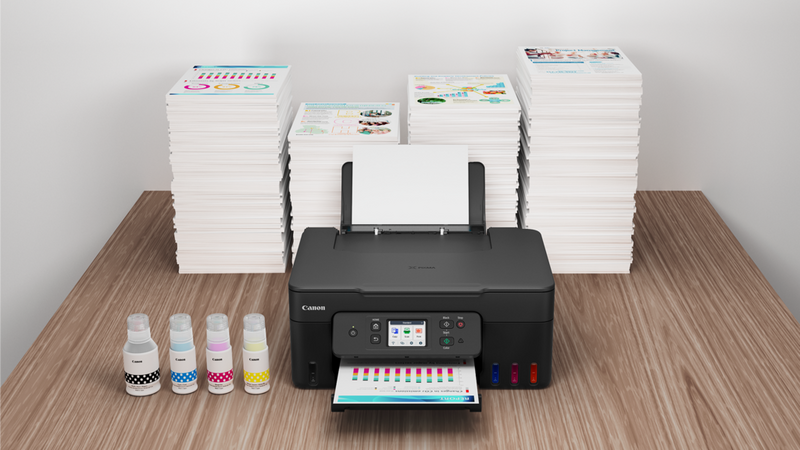 Printer fra PIXMA G-serien (MegaTank) på et skrivebord med blækflasker, papirstakke og et farveprint, der kommer ud.
