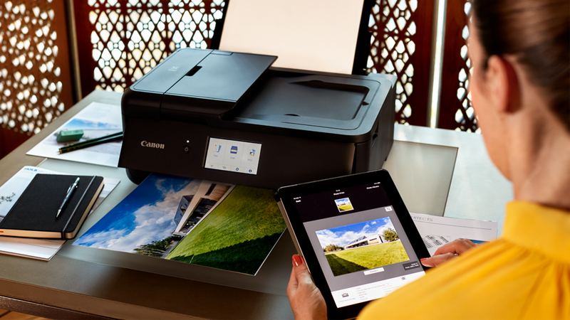 Canon PRINT-appen på en tablet ved siden af en Canon-printer, der printer et foto på et skrivebord.