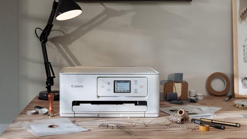 Printer i PIXMA TS-serien på et skrivebord med brevpapir, papir og en lampe på en hjemmearbejdsplads.