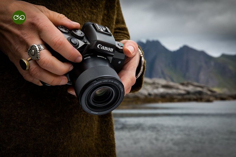 Person holding a Canon mirrorless camera beside a lake with mountains in the background