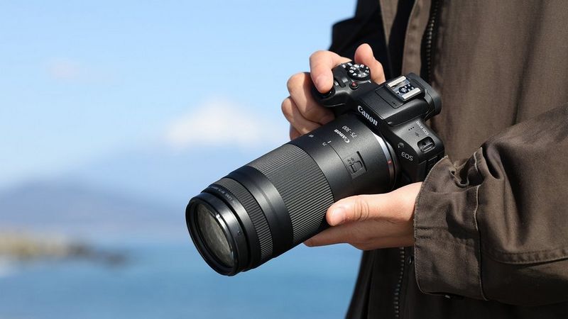 Persona sosteniendo una cámara mirrorless Canon EOS R100 con un objetivo RF 75-300mm frente a un cielo azul