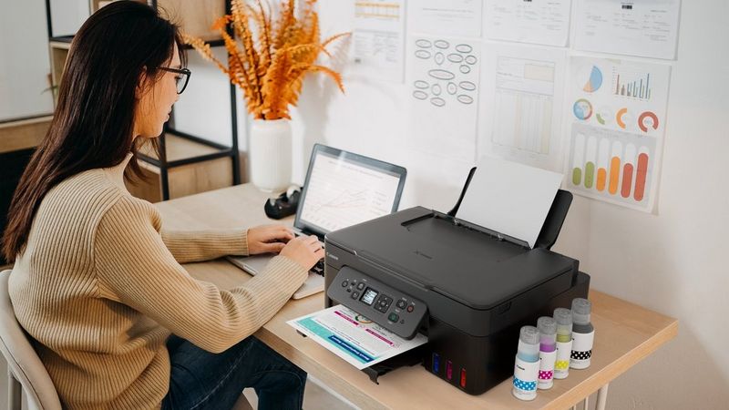 Person using a laptop beside a Canon printer printing a colourful document in a home office workspace