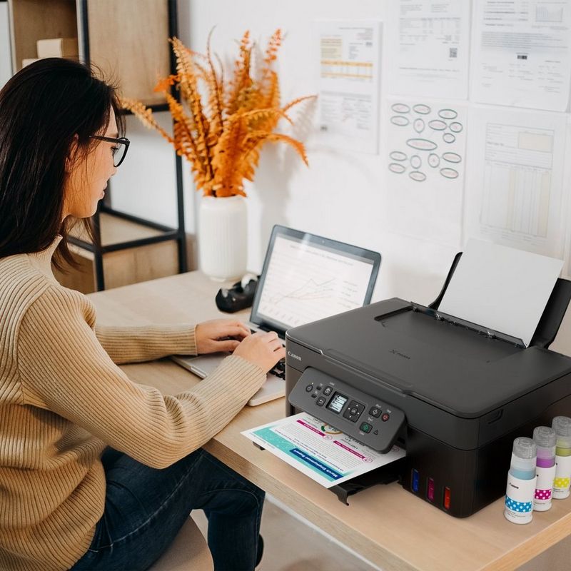 Person using a laptop beside a Canon printer printing a colourful document in a home office workspace