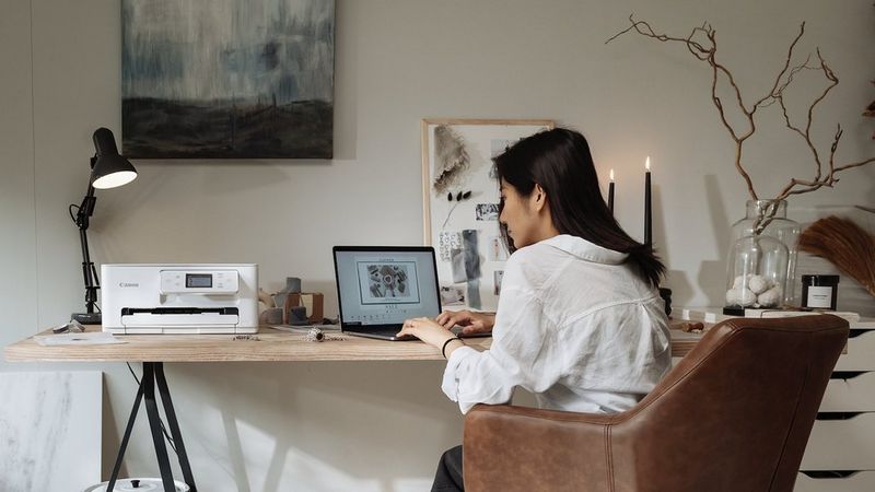 Person working on a laptop beside a Canon printer in a home office with artwork and desk décor.