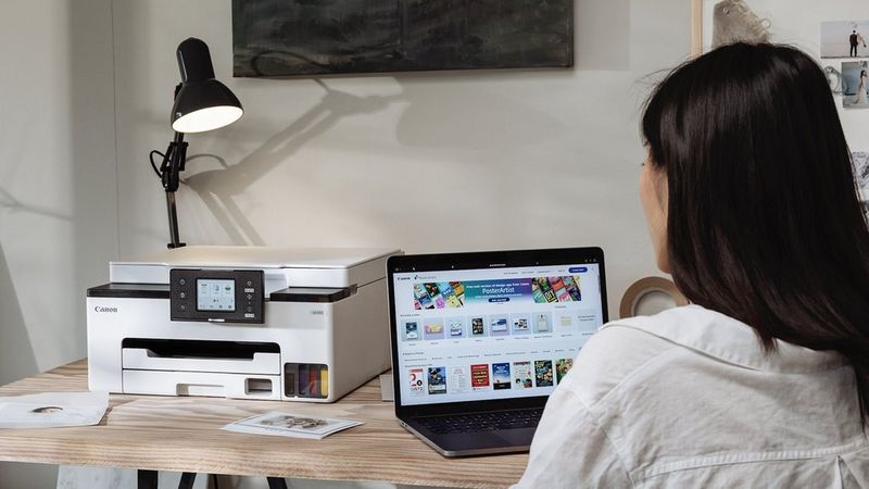 Person using a Canon MAXIFY GX1015 Printer with a laptop in a home office workspace.