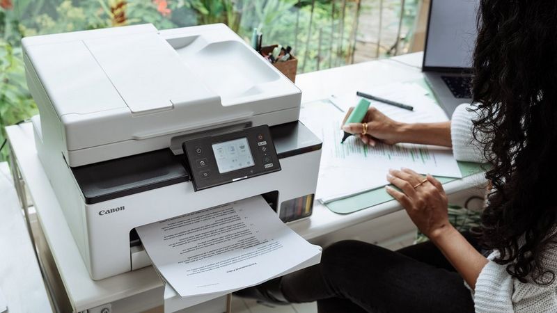 Canon MAXIFY GX2051 printer beside a person writing in a notebook in a home office.
