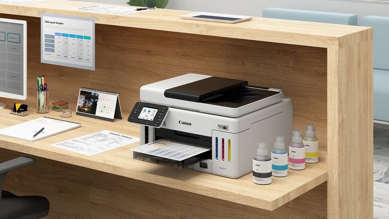 Canon MAXIFY GX6150 printer on a wooden office desk printing documents beside ink bottles and stationery.