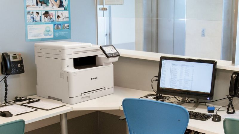 Canon i-SENSYS MF664Cdw printer on an office desk beside a computer monitor.