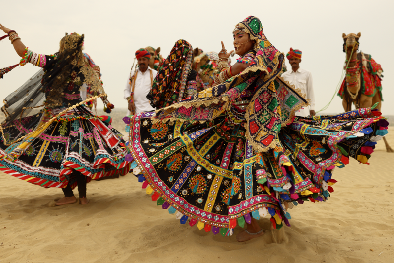 Gruppe von Menschen in bunten Kleidern, die auf Sand tanzen und hinter der Kamele zu sehen sind.