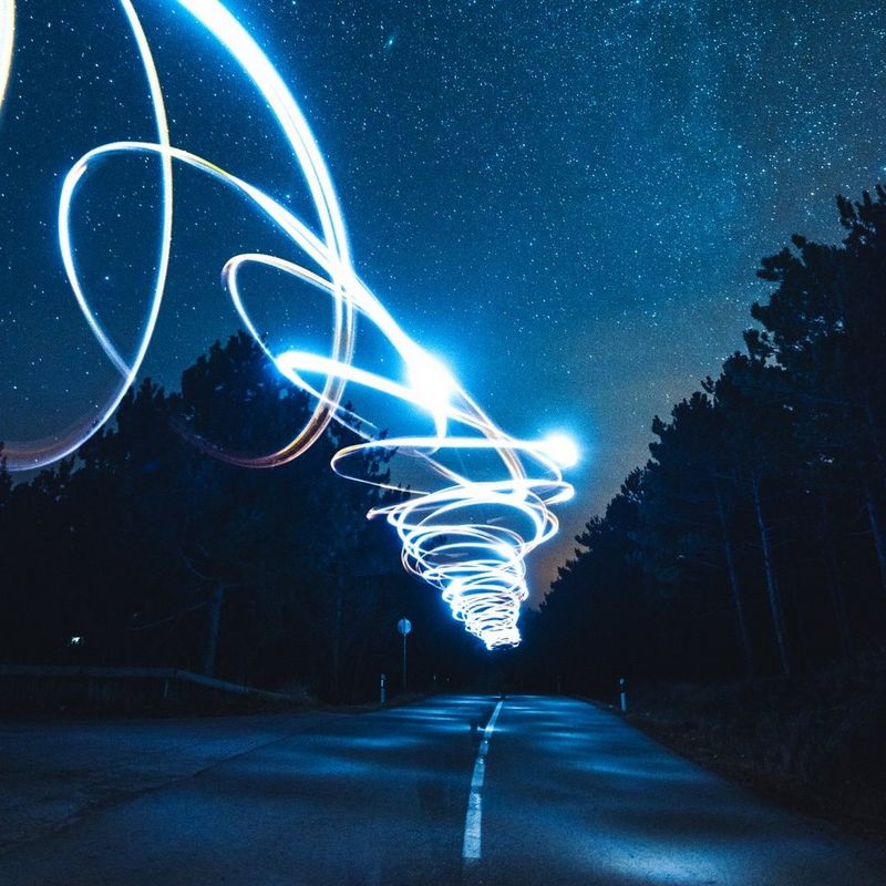 Blue and white light trails spiral above a forest road beneath a star-filled night sky.