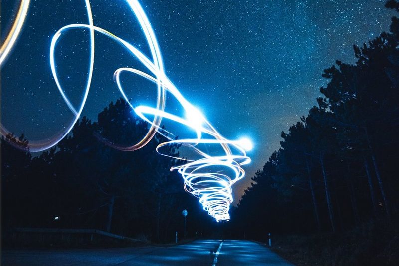 Blue and white light trails spiral above a forest road beneath a star-filled night sky.