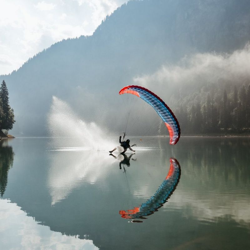 Een paraglider vliegt over een rustig bergmeer, met waterdruppels en een kleurrijke parachute weerspiegeld in het water eronder