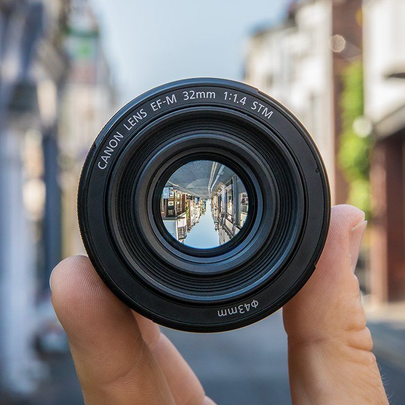 A hand holding lens EF-M 32MM F1.4 STM in the street showing the same street upside down through the lens.