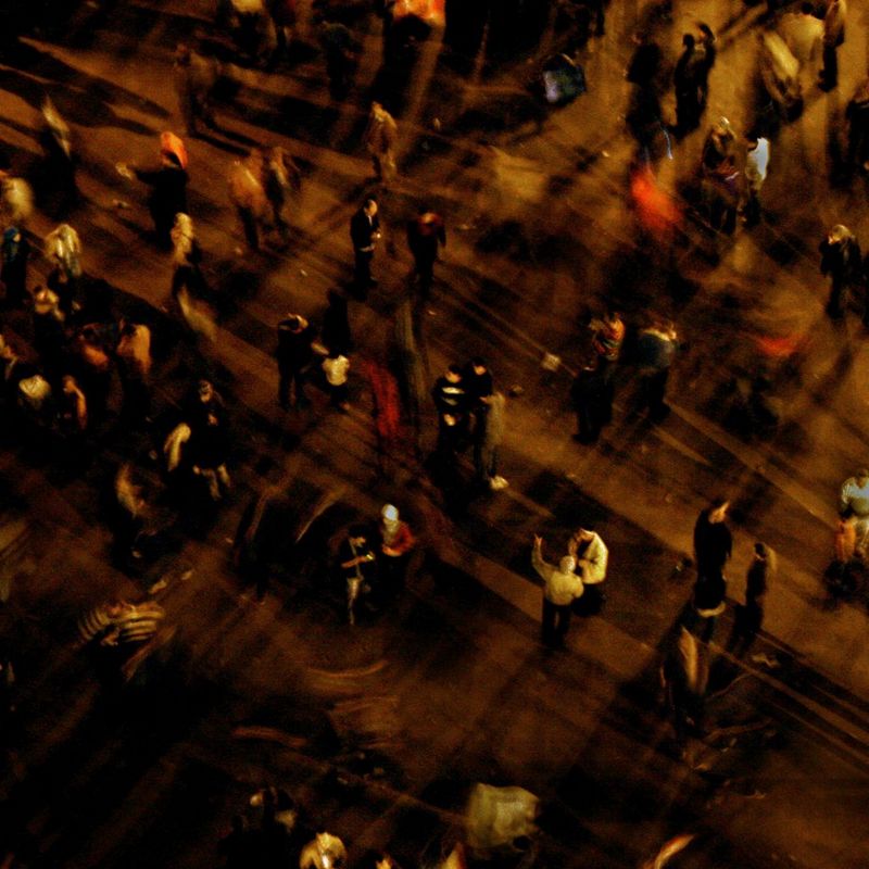 A top-down image showing groups of people standing in a square. A long exposure has blurred the figures who cast long shadows on the ground.