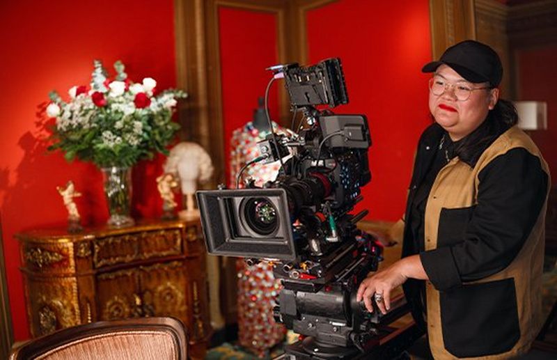 A woman wearing a cap and glasses, dressed in a black and tan jacket, stands behind a Canon D224 setup in a luxurious room.