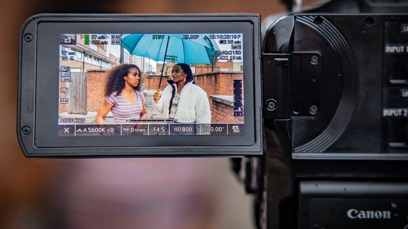 A close-up of the LCD screen of a Canon EOS C70 showing two women talking under a large blue umbrella, as well as numerous camera settings.