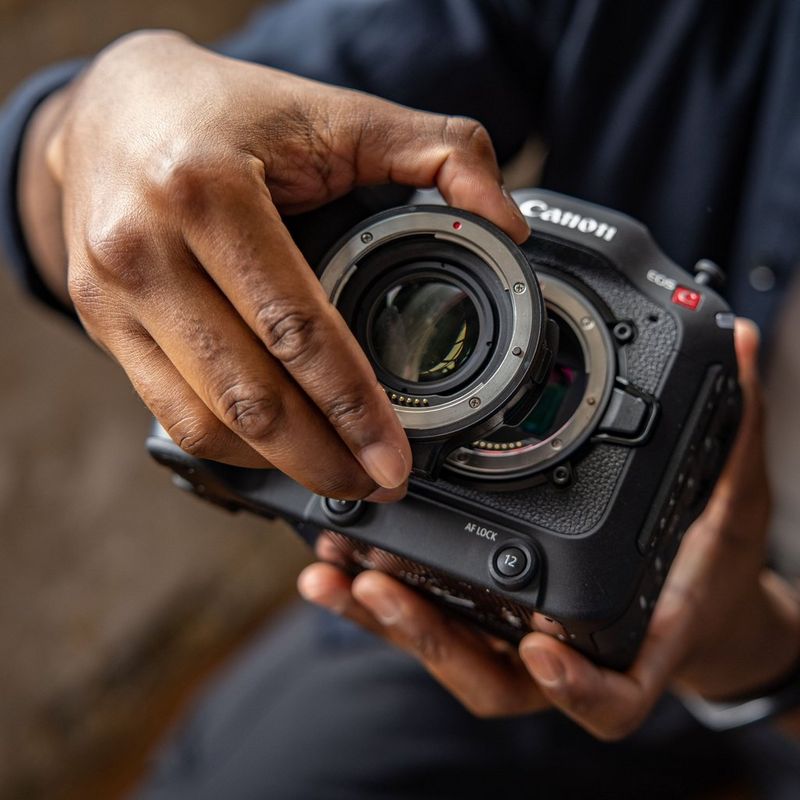 A close-up of a user's hands fitting a Canon Mount Adapter EF-EOS R 0.71x to a Canon EOS C70 camera.
