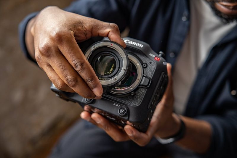 A close-up of someone's hands fixing the Mount Adapter EF-EOS R 0.71x to a Canon EOS C70 camera. 