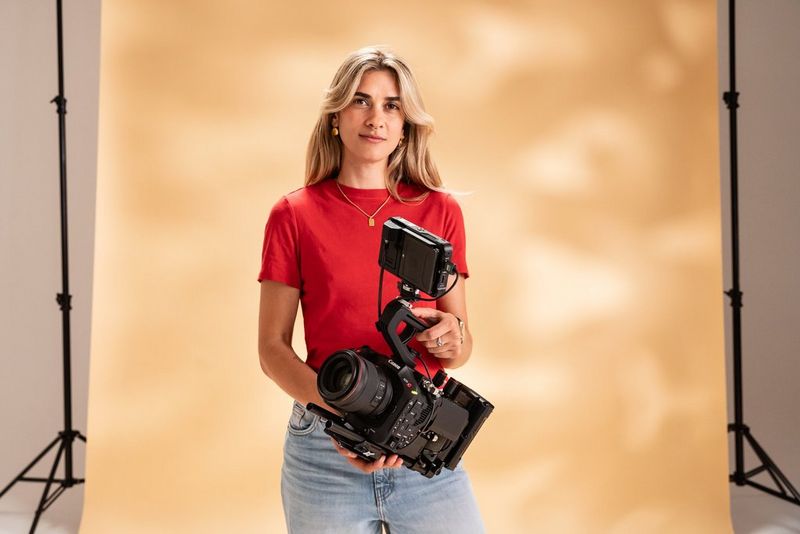 Cinematographer Carolina Nunes, in a red top and jeans, standing in front of a creamy-coloured backdrop holding the Canon EOS C80 camera.