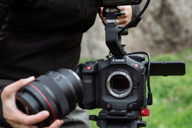A user holds a Canon lens in one hand, ready to attach it to a Canon EOS C80 with no lens attached, showing the RF mount and sensor.
