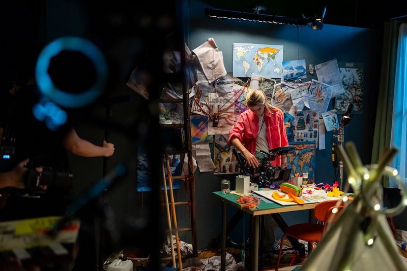 A cinematographer, Carolina Nunes, holds a Canon EOS C80 in her hands to film a table of colourful objects. She is standing in front of a blue wall covered in paper cuttings. 