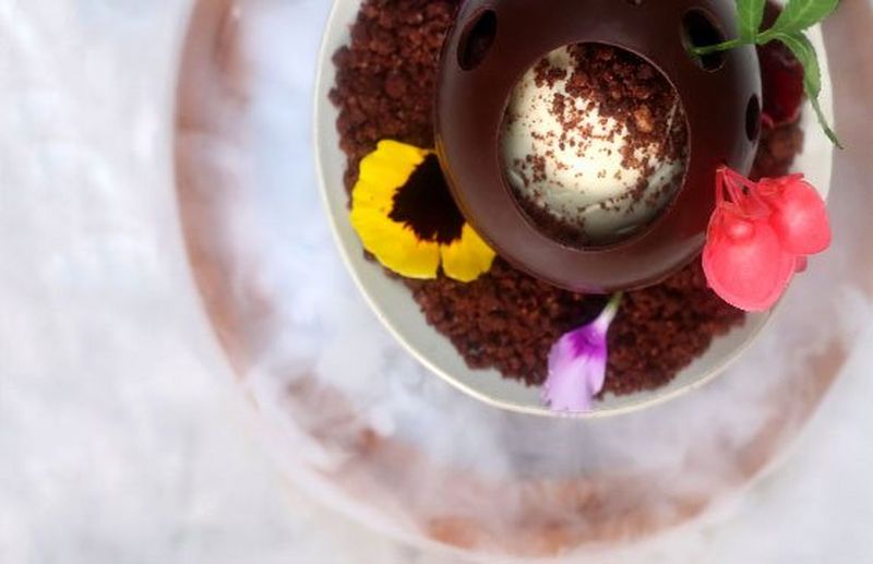 A hollow chocolate ball filled with ice cream and garnished with petals and chocolate sprinkles. 