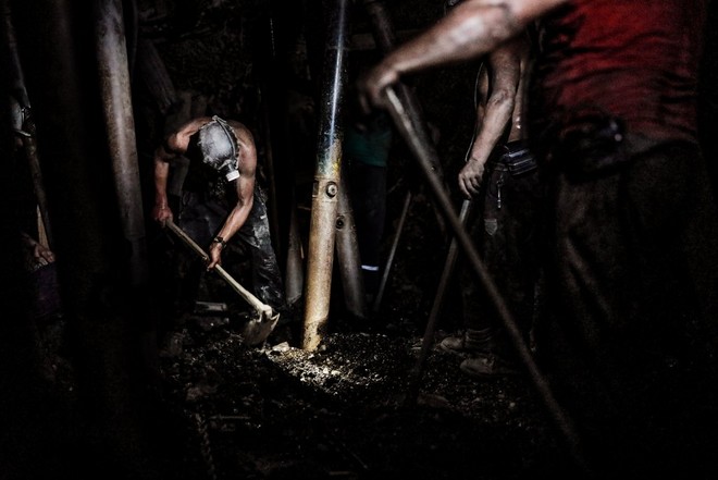 Um mineiro a trabalhar no solo de uma mina pouco iluminada, com dois outros mineiros a olhar.