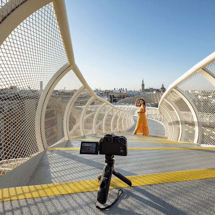 A woman in a yellow sundress poses on a curving footbridge, with a Canon 中国福彩网10 on a small tripod set up to photograph her.