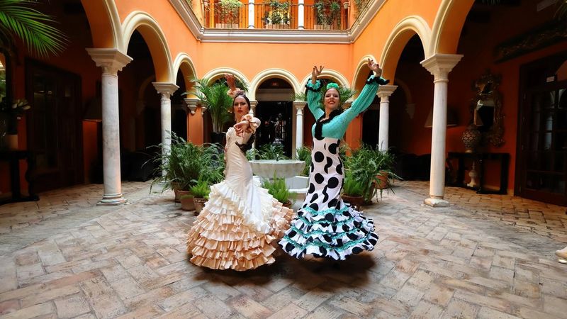 Două dansatoare de flamenco în rochii tradiţionale pozează cu braţele ridicate într-o curte.