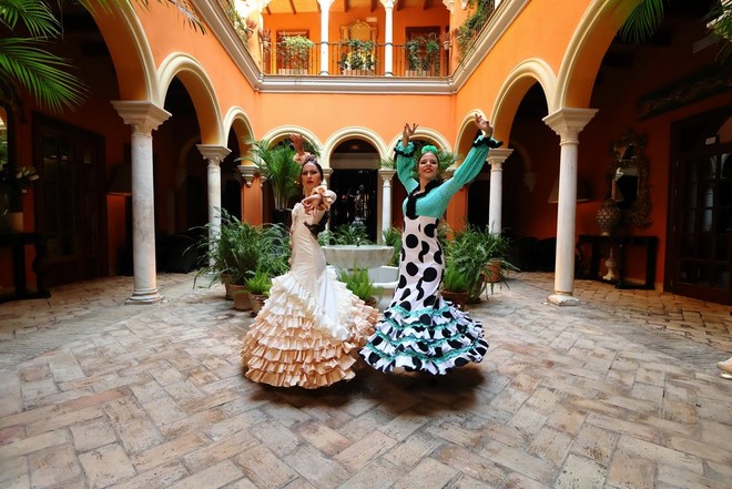 Duas dançarinas de Flamenco com vestidos compridos com folhos a posar para a câmara num pátio colorido com vários arcos.