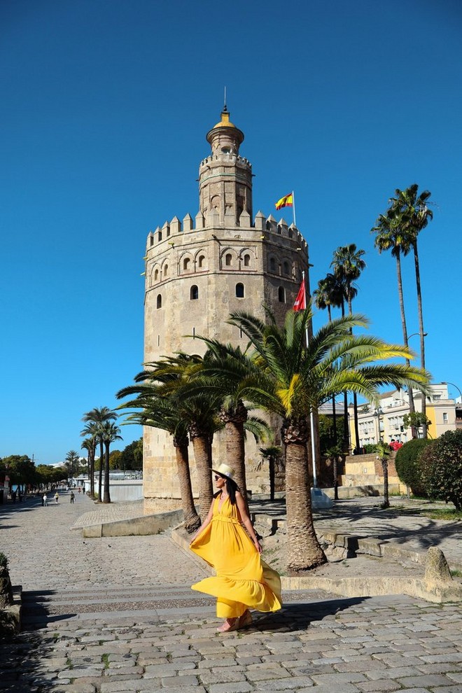 امرأة ترتدي فستانًا أصفر تسير في ممر تمتد على جانبه أشجار النخيل بينما يظهر برج حجري في الخلفية.