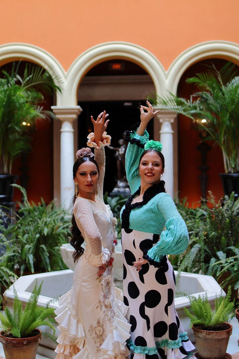 Two flamenco dancers in a traditional dress pose with arms raised and hands on their hips in a leafy courtyard.