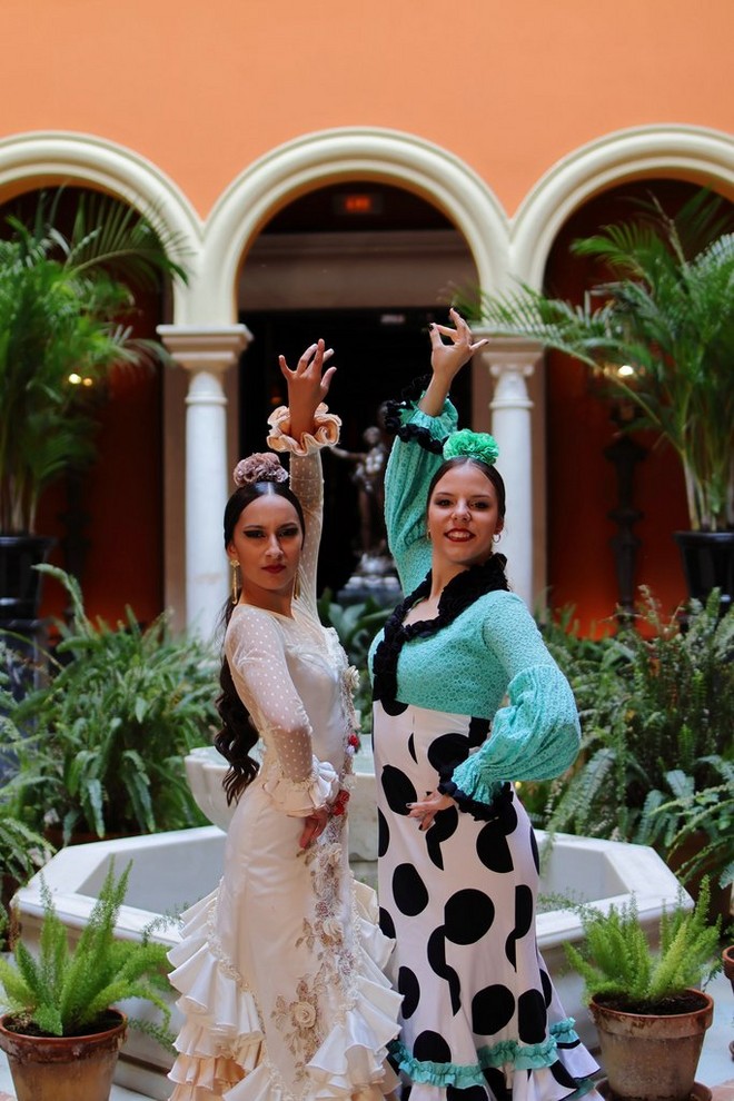 Two flamenco dancers in a traditional dress pose with arms raised and hands on their hips in a leafy courtyard.