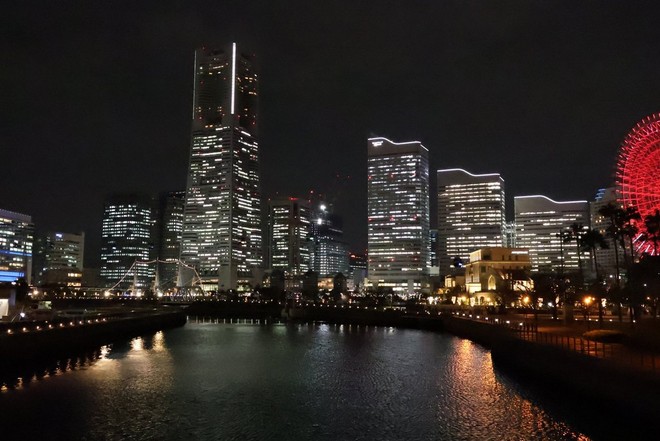 A night-time cityscape showing tall buildings with street lights reflected in a body of water in the foreground. 