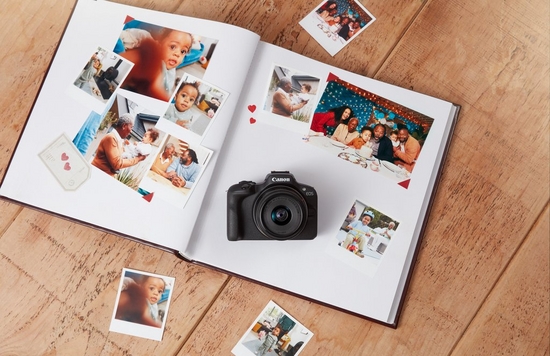 A Canon EOS R100 camera lies on a photo album on a wooden table, surrounded by family photographs.
