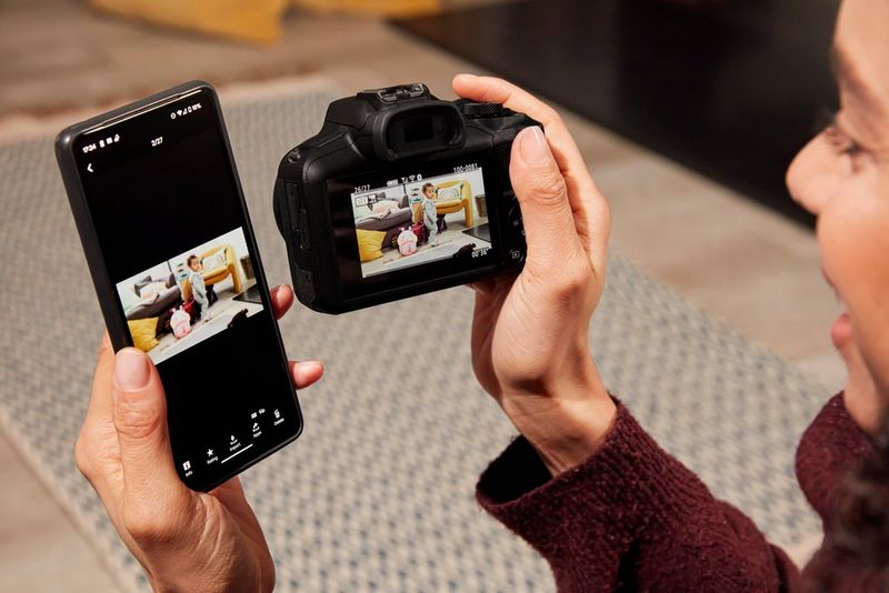 A figure holds a Canon EOS R100 camera next to a smartphone, the same image being displayed on the camera screen and Canon Camera Connect app on the phone.