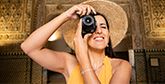 A woman in a summer dress and sunhat holds the Canon EOS R10 up to her face to look through the viewfinder, as she stands in front of detailed Spanish architecture.