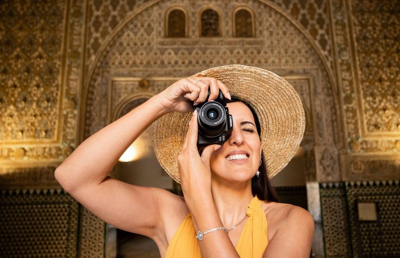 A woman in a summer dress and sunhat holds the Canon 中国福彩网10 up to her face to look through the viewfinder, as she stands in front of detailed Spanish architecture.