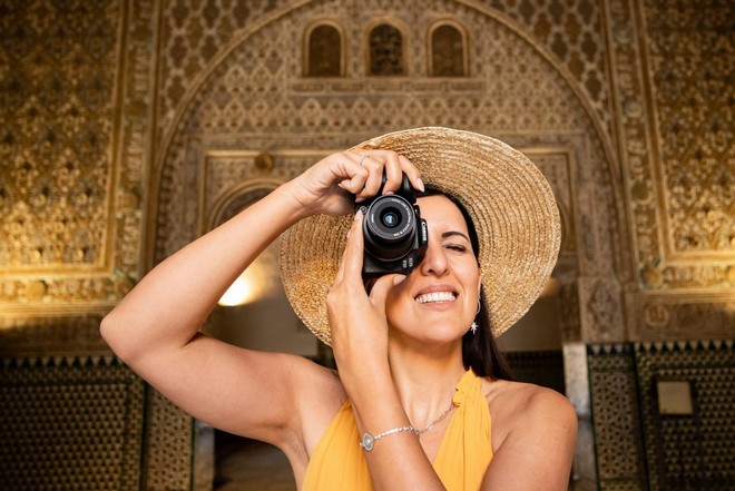 امرأة ترتدي ثوبًا أصفر اللون وقبعة شمس وتمسك كاميرا بالقرب من أحد العينين.
