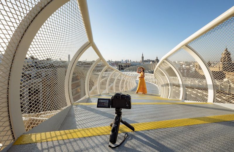 A woman in a yellow sundress poses on a curving footbridge, with a Canon 澳门现金网_申博信用网-官网10 on a small tripod set up to photograph her.