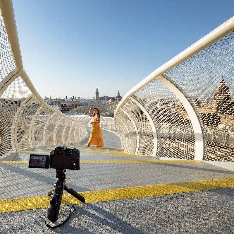 Un Canon EOS R10 monté sur un trépied au premier plan pointe vers une femme à mi-distance sur une passerelle incurvée, avec une ville à l'arrière-plan.