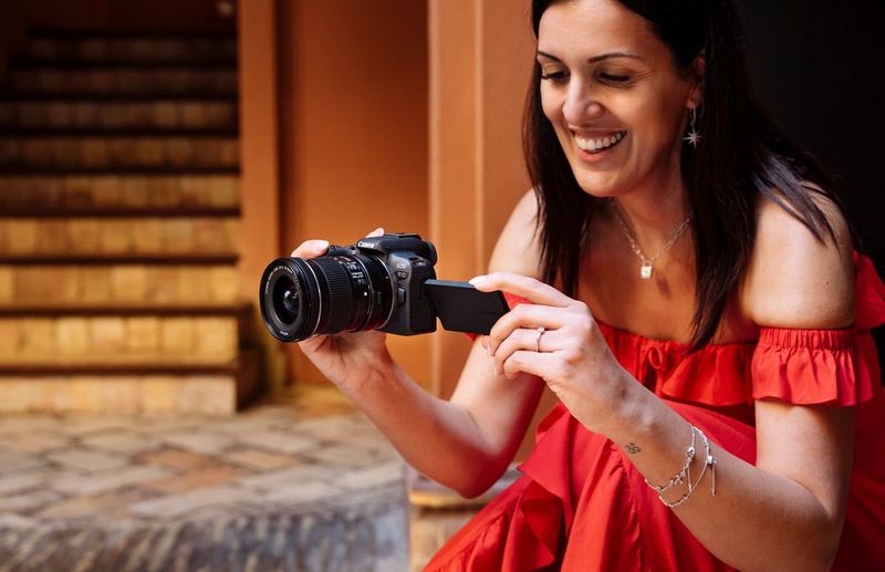 A woman wearing a red strapless dress crouches down to take a picture with a Canon EOS R10.